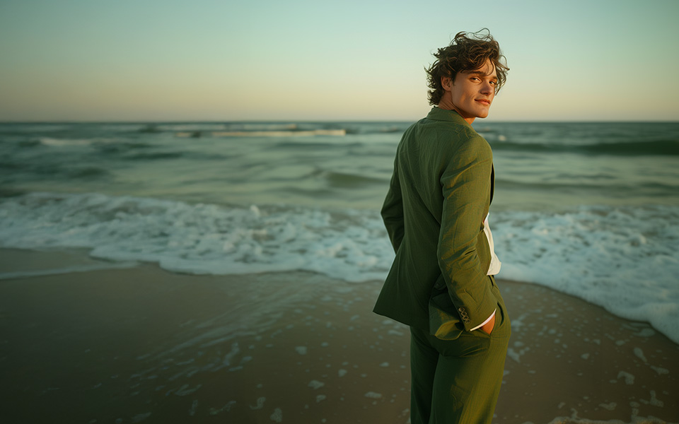 KI-generiertes Bild eines jungen Mannes bei Abenddämmerung am Strand. Er trägt einen olivgrünen Leinenanzug.