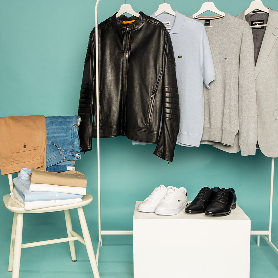 Verschiedene Kleidungsstücke wie Lederjacke, Sakko, Pullover und Polo-Shirt hängen an einer Garderobe vor türkisfarbener Wand. Davor stehen weiße Sneaker und schwarze Businessschuhe auf einem Podest. Auf einem Stuhl daneben liegen gefaltete T-Shirts und Hemden in Weiß, Beige und Hellblau; über der Lehne hängen eine Jeans und eine beige Chino.