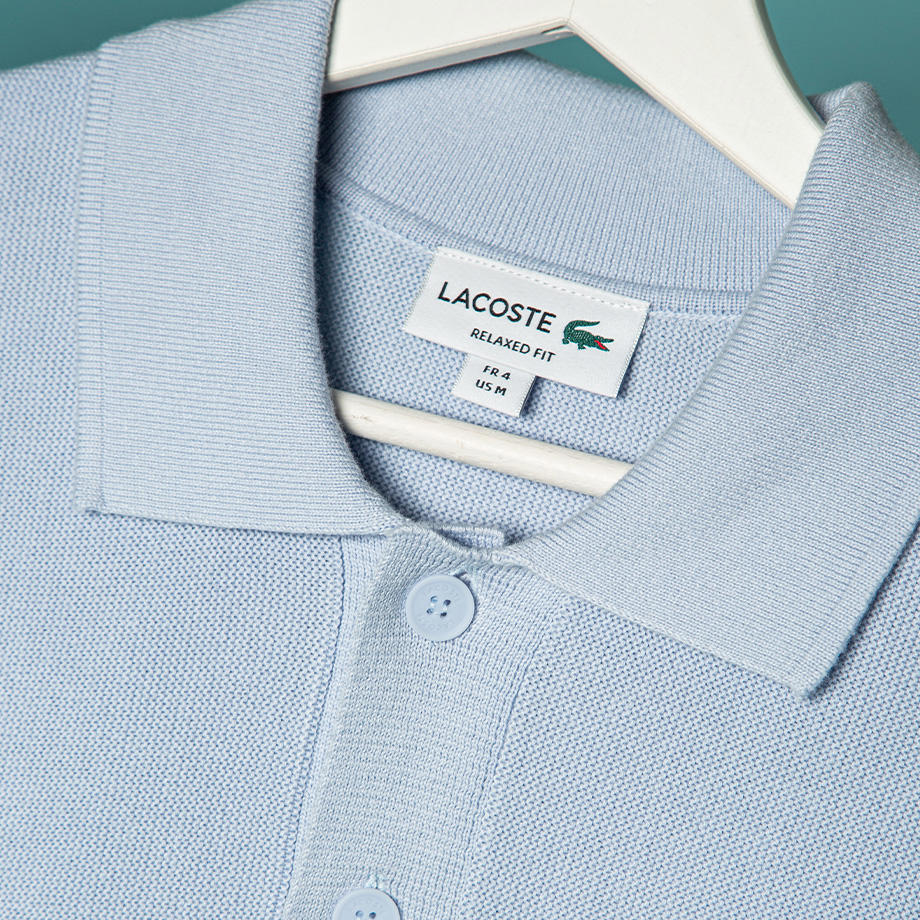 Detailaufnahme eines hellblaues Poloshirt von Lacoste auf einem weißen Kleiderbügel vor grünem Hintergrund.