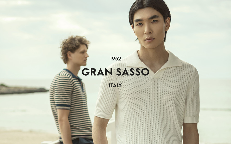 Zwei junge Männer stehen auf einem Holzsteg am Strand. Sie tragen sommerliche Looks. In der Mitte ist das Logo: Gran Sasso Italy