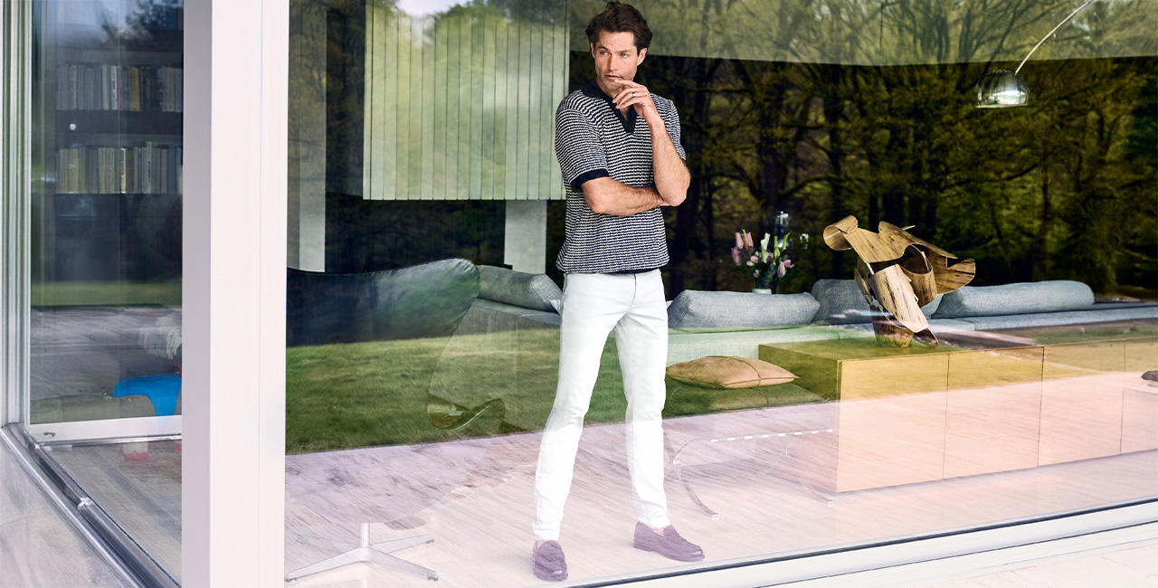 Ein Mann in weißen Hosen von Alberto und schwarz-weiß gestreiftem Polo-Shirt steht an einer Fensterfront und blickt nach draußen.