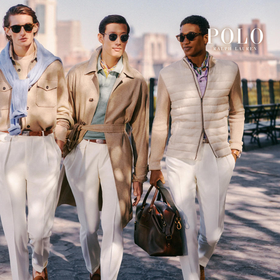Drei stilvoll gekleidete Männer gehen nebeneinander auf einem sonnigen Gehweg in der Stadt. Sie tragen helle, elegante Outfits in Beige- und Pastelltönen, darunter weiße Bundfalten Hosen, Pullover, beige Mäntel und Jacken. Alle drei tragen Sonnenbrillen. Im Hintergrund sind Hochhäuser zu sehen. Oben rechts im Bild steht das Logo „POLO RALPH LAUREN“
