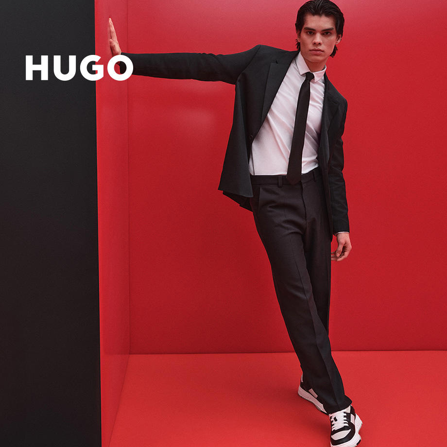 Ein junger Mann posiert in einem leuchtend roten Raum, an die Wand gelehnt. Er trägt einen schwarzen Anzug, weißes Hemd, schwarze Krawatte und Sneakers. Der Markenname „HUGO“ ist über das Bild gelegt.