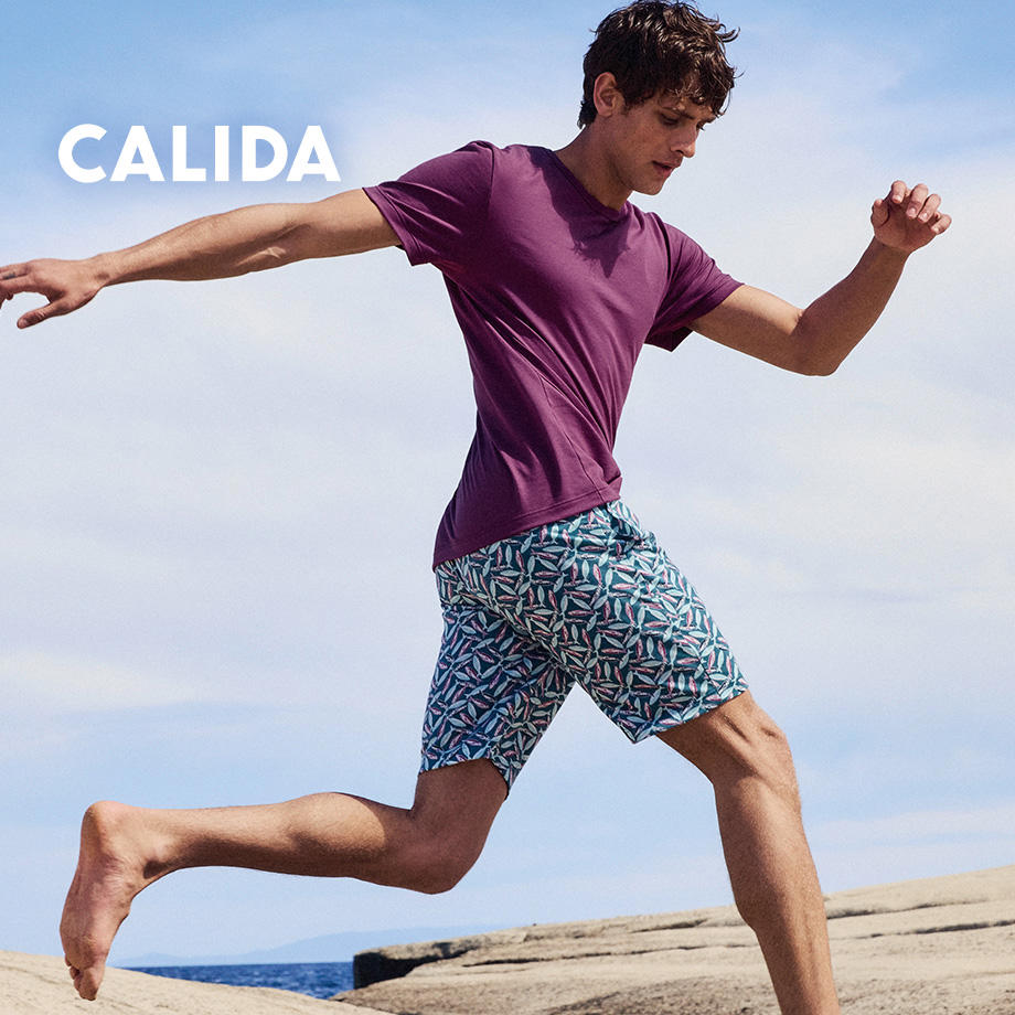 Sportlicher Mann in violettem T-Shirt und gemusterten Shorts von Calida, springt am Meer – bequeme Bademode für aktive Männer.