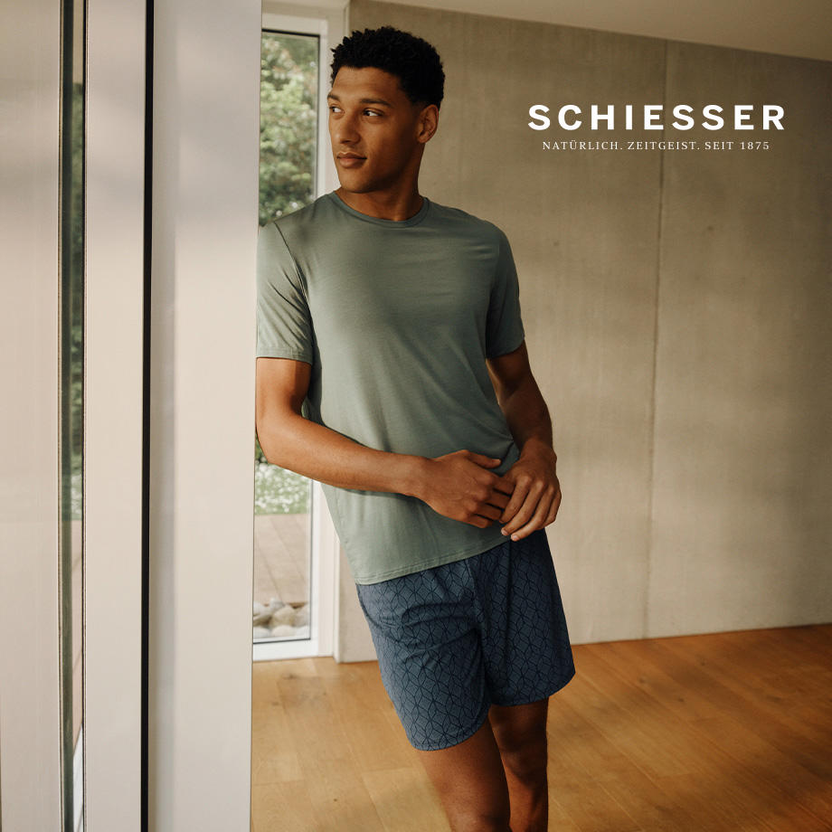 Mann trägt grünes T-Shirt und blaue Shorts von Schiesser, steht entspannt am Fenster – moderne Herrenloungewear mit Sommerflair.