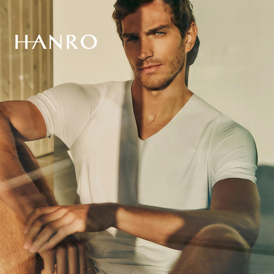 Attraktiver Mann in weißem V-Ausschnitt-Shirt von Hanro, sitzt auf Sofa – elegante Herrenunterwäsche in Premiumqualität