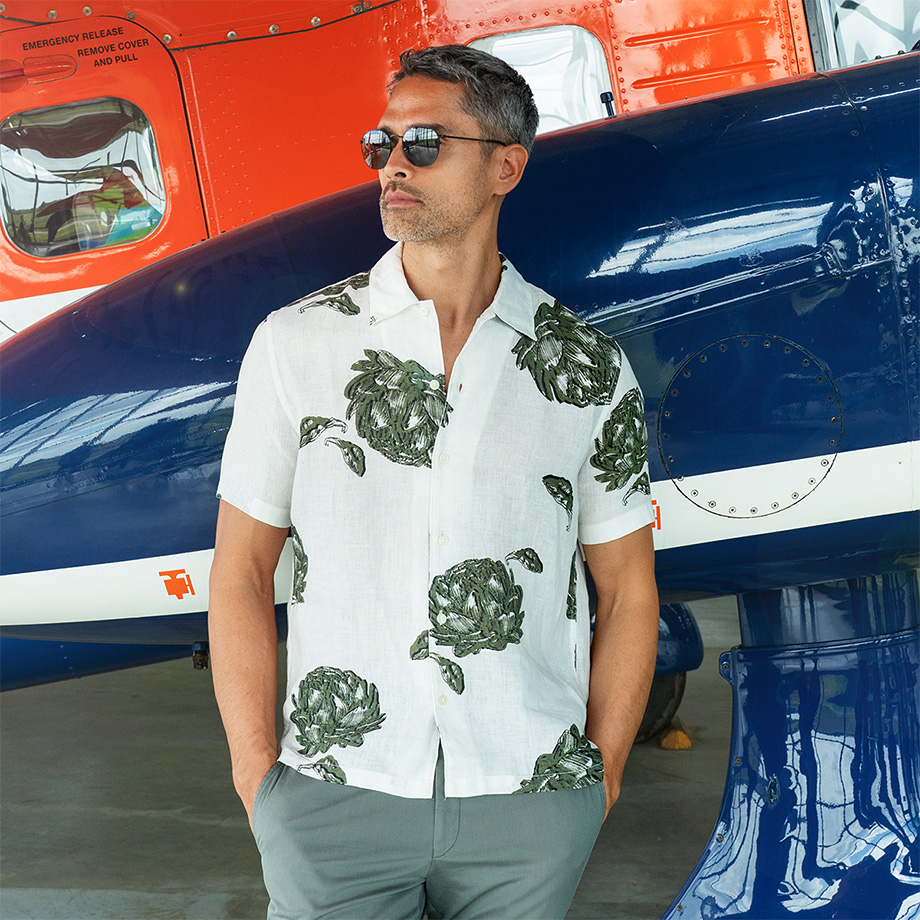 Mann mit Sonnenbrille in sommerlichem Freizeit-Outfit: weißes Kurzarmhemd mit großem grünen Pflanzenmuster und grauer Hose. Er steht vor einem farbenfrohen Flugzeug – stilvoller Urlaubs-Look für warme Tage.