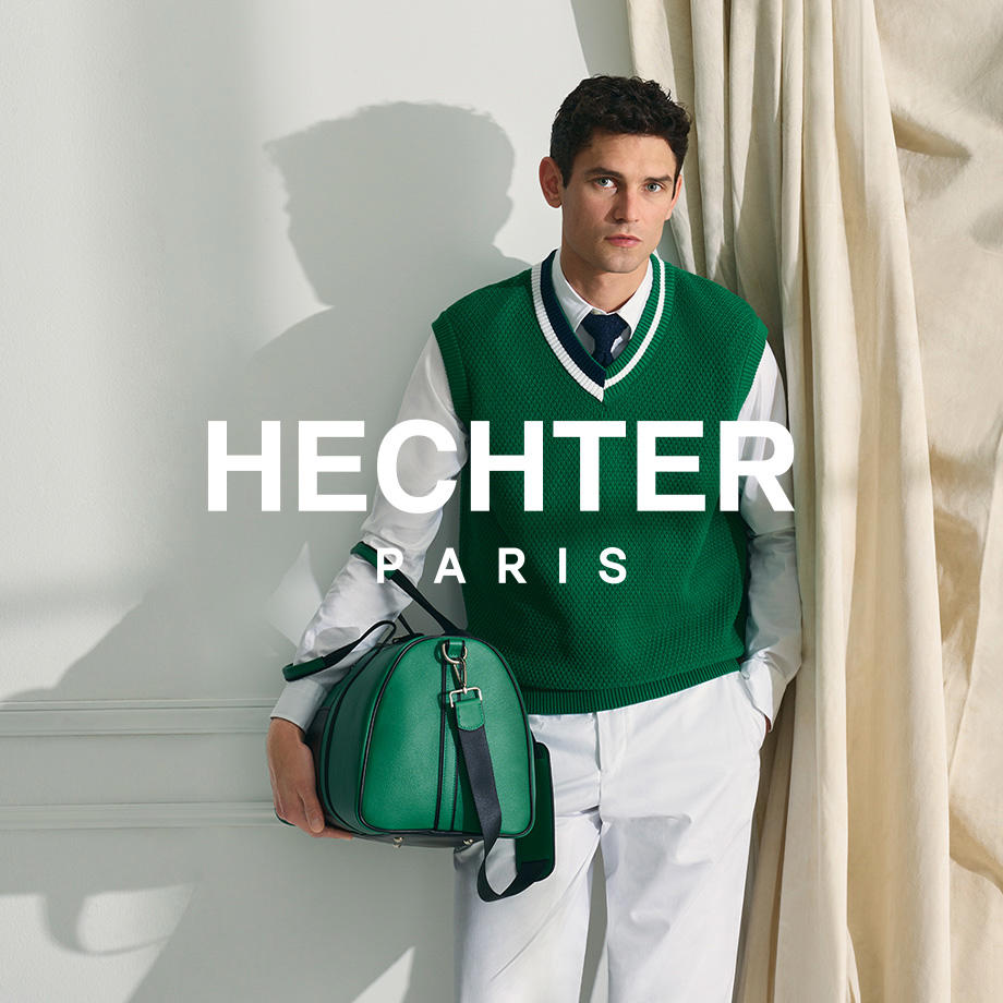 Ein Mann steht vor einem hellen Vorhang in elegantem Outfit von Hechter Paris: Er trägt ein weißes Hemd mit grünem Strickpullover, weiße Hose und hält eine grüne Weekender-Tasche – ein stilvoller, moderner Look mit französischem Flair.