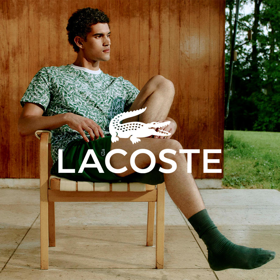Ein Mann sitzt bequem in einem Stuhl vor einer hellen Holzwand auf der Terasse im Garten. Er trägt ein lässiges grünes Shirt eine Short und Socken.