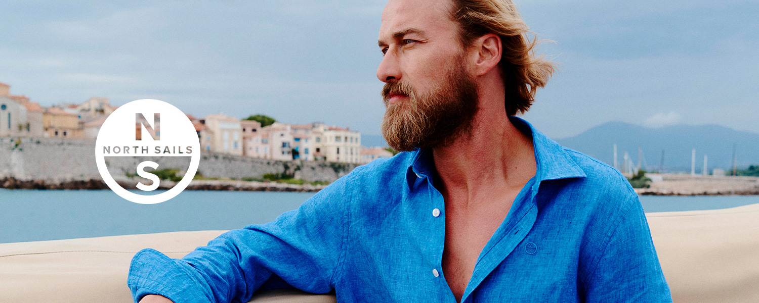 Mann in offenem blauen Jeanshemd lehnt lässig an einer Bootsreling mit Blick auf eine mediterrane Stadt.