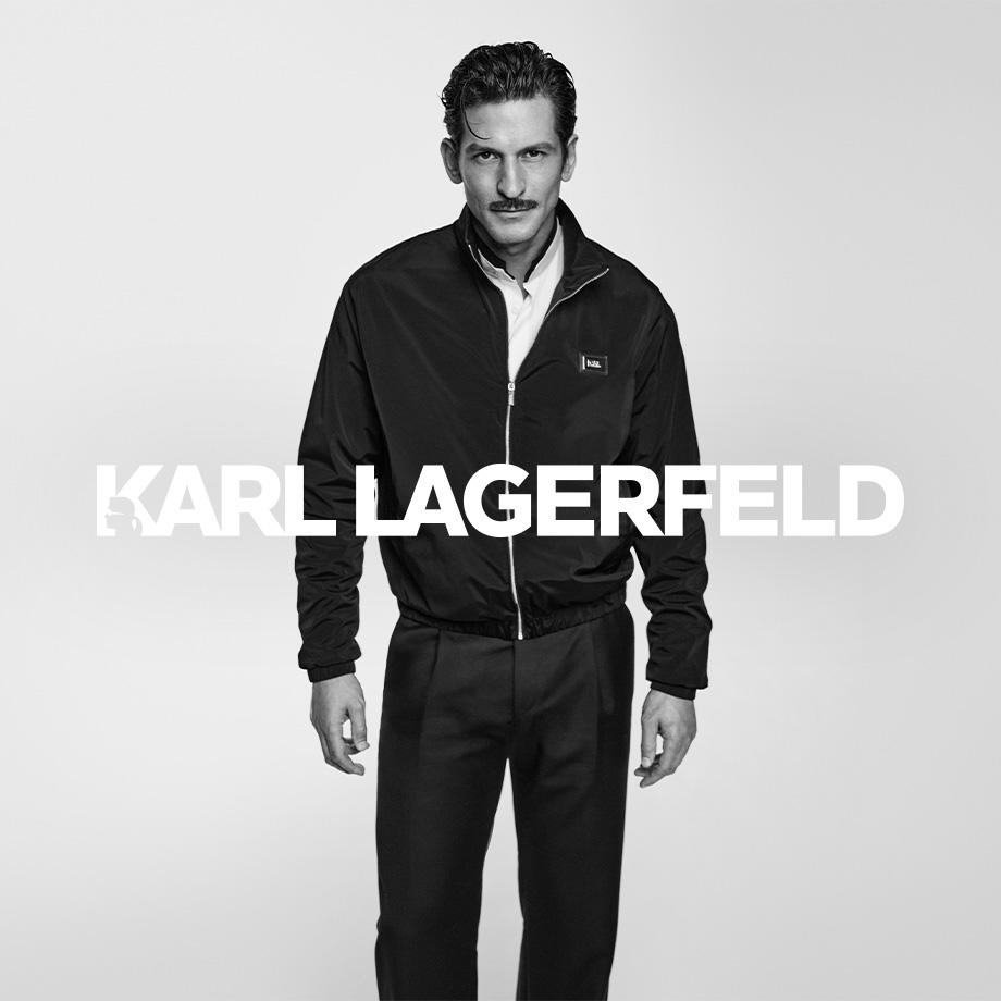 Ein Mann steht vor neutralem Hintergrund in einem minimalistischen Schwarz-Weiß-Porträt. Er trägt ein elegantes, sportlich inspiriertes Outfit von KARL LAGERFELD mit dunkler Jacke, weißem Hemd und dunkler Hose – ein moderner, markanter Look mit ikonischem Stil.