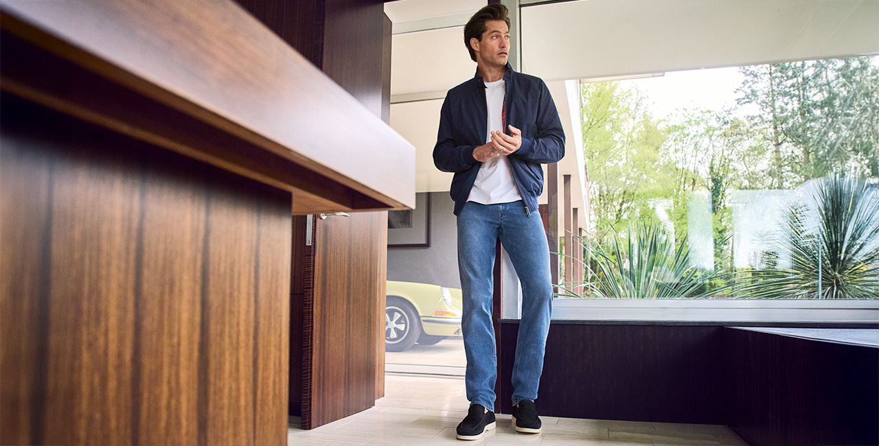 Mann in Jeans, weißem T-Shirt und dunkelblauer Jacke steht in einem modernen, lichtdurchfluteten Innenraum mit Holzverkleidung und Blick auf einen Garten.
