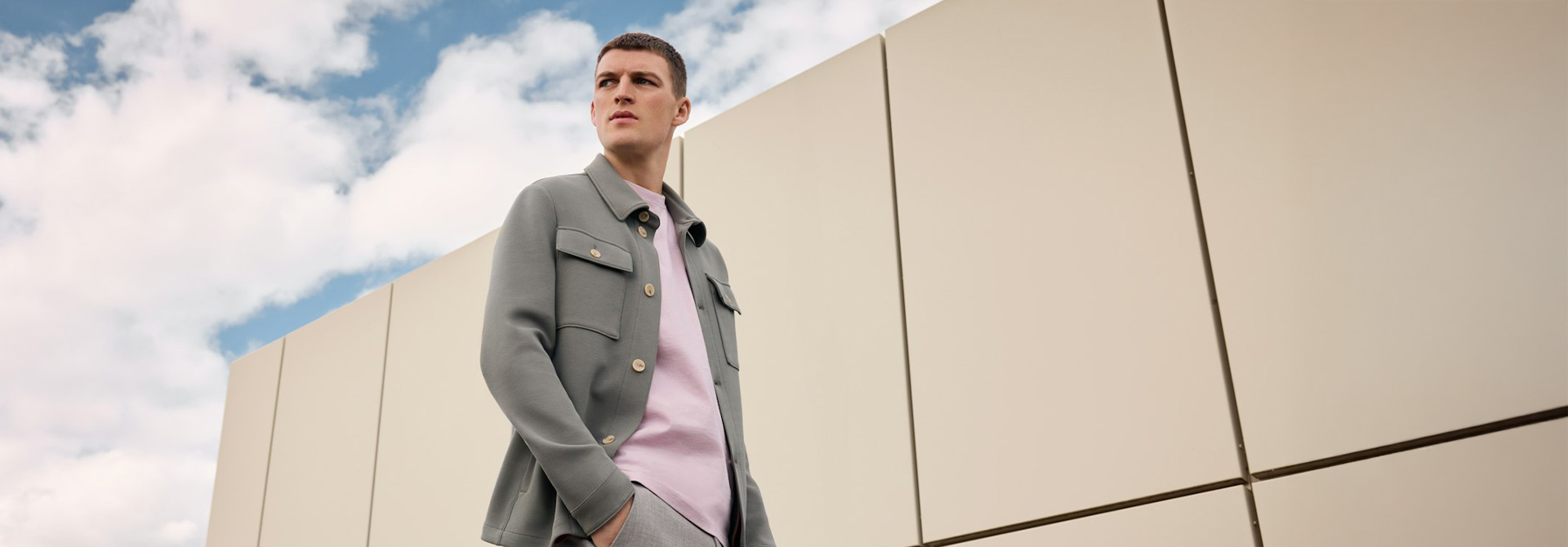 Ein junger Mann steht vor einem weißen Gebäude. Er trägt ein olivgrünes Overshirt.