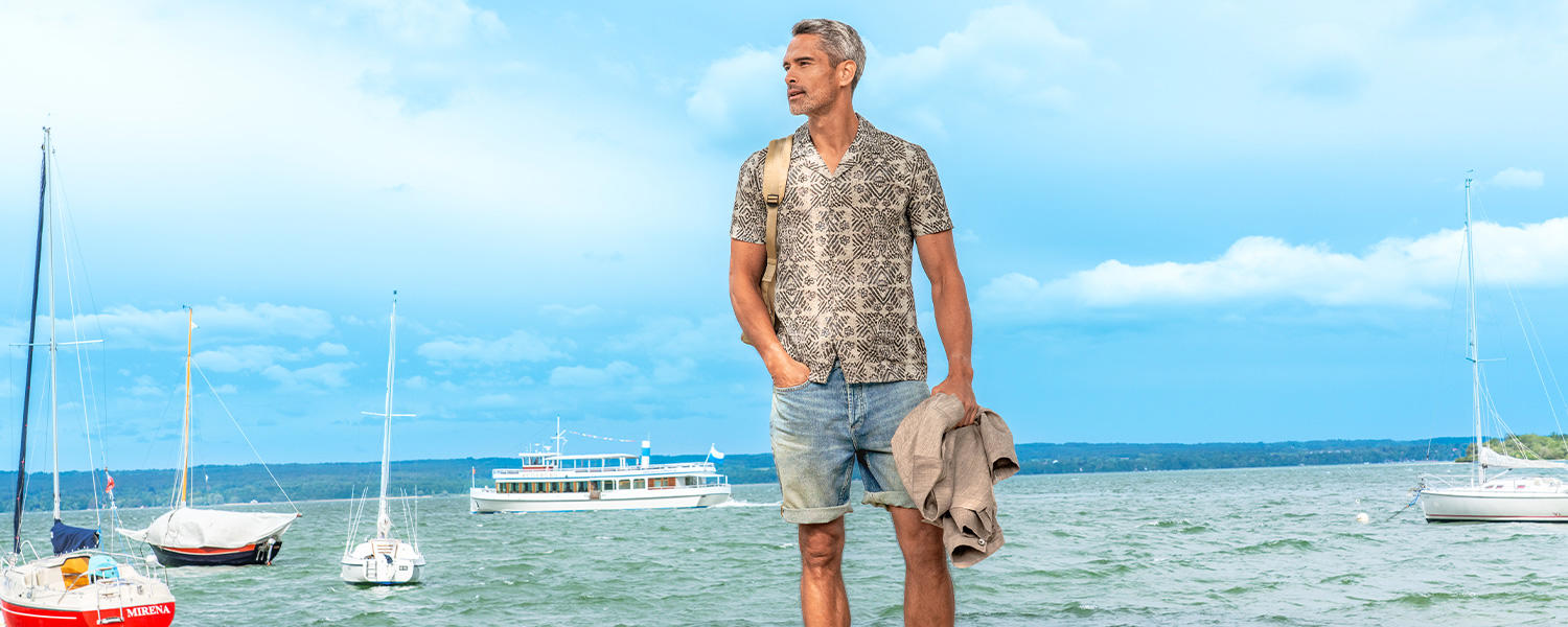 Ein Mann mittleren Alters,  mit leichten grauen Haaren steht entspannt am Ufer eines Sees. Er trägt ein gemustertes, kurzärmliges Hemd, hellblaue Jeans-Shorts und hat eine beigefarbene Jacke in der Hand sowie einen Rucksack über der Schulter. Im Hintergrund sind Segelboote und ein Ausflugsschiff auf dem Wasser zu sehen, unter einem leicht bewölkten, blauen Himmel.