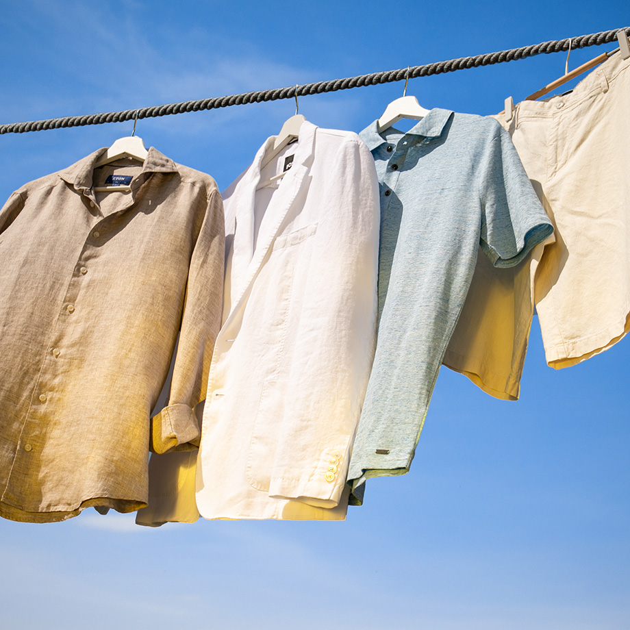 Leinenhemden, ein Leinen Polo-Shirt und eine Leinenhose in Naturtönen hängen auf einer Wäscheleine vor blauem Himmel.