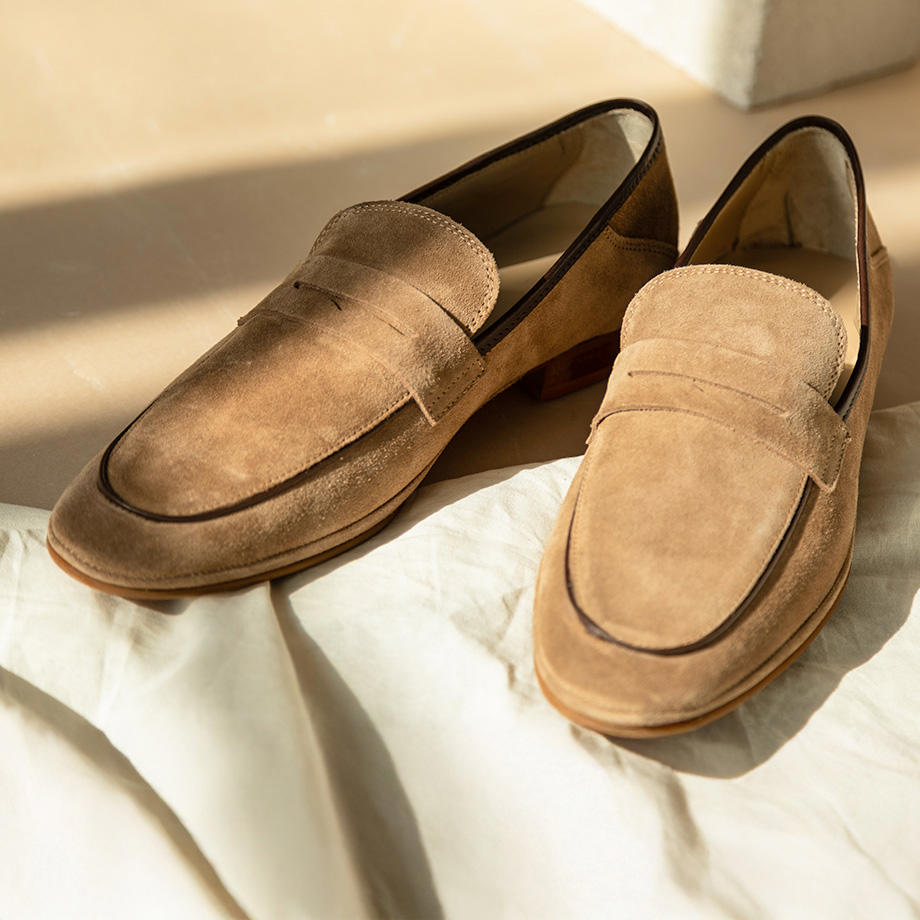 Klassische beigefarbene Loafer aus Wildleder auf heller Stoffunterlage.