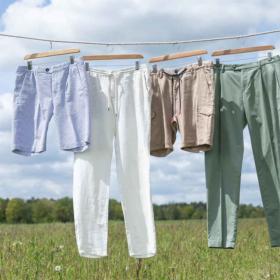 Sommerliche Chinohosen und Shorts in hellen Farben hängen auf einer Leine über einer grünen Wiese vor blauem Himmel.
