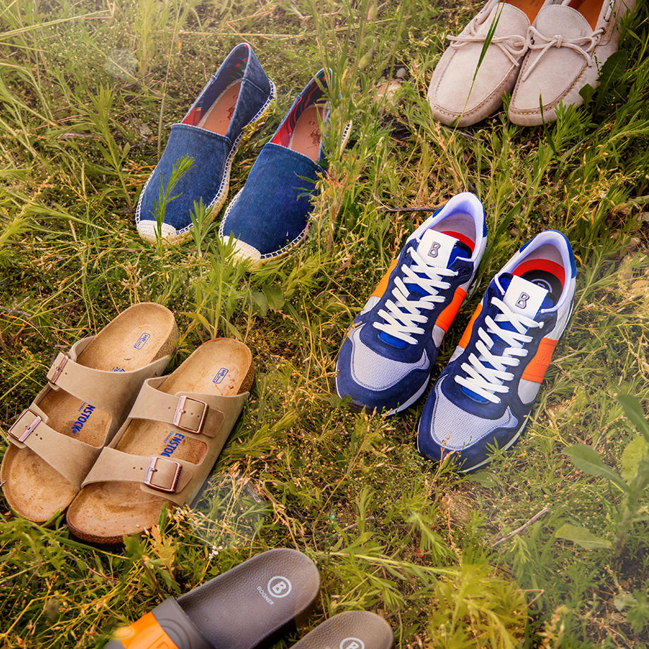 Verschiedene Herrenschuhe, darunter Sandalen, Sneaker, Loafer und Espadrilles, liegen auf einer Wiese im Sonnenschein.