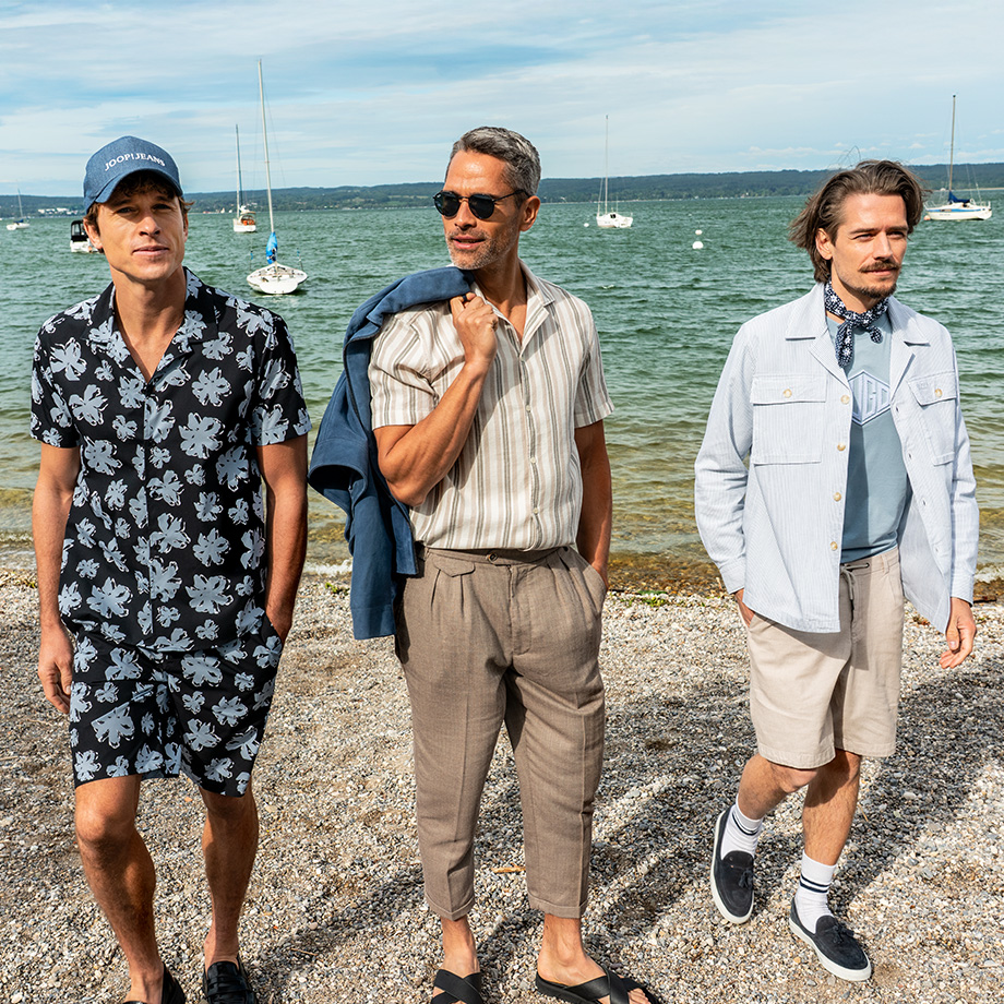 Drei Männer stehen am Ufer eines Sees mit Segelbooten im Hintergrund. Sie tragen sommerliche Outfits in gedeckten Tönen: links ein Ensemble mit floraler Hemd-Shorts-Kombination in hell- und dunkelblau und blauer Cap, in der Mitte ein gestreiftes Hemd mit beiger Stoffhose und blauer Lederjacken über der Schulter, rechts ein heller Look mit beigen Shorts, hellblauem T-Shirt, Jacke und Halstuch.