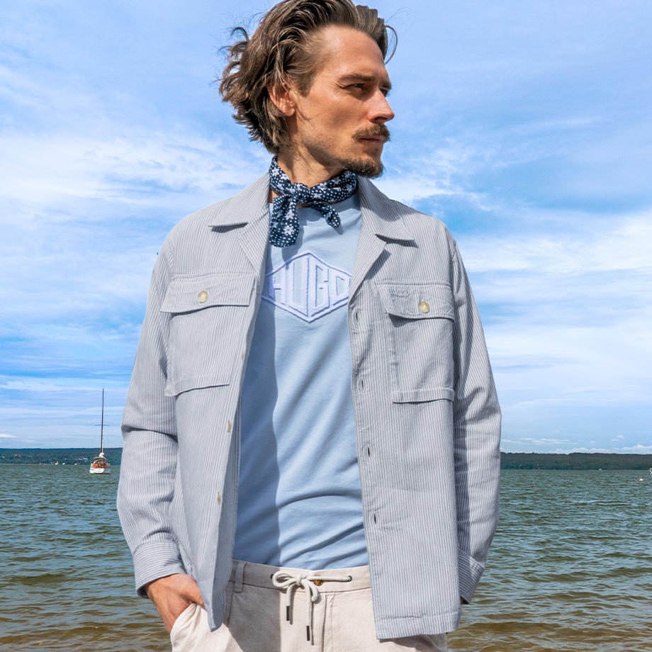 Ein Mann steht vor einer Hauswand mit Doppeltüren. Er trägt ein helles Hemd, beigefarbene Shorts und weiße Sneaker – ein stilvoller Sommerlook.