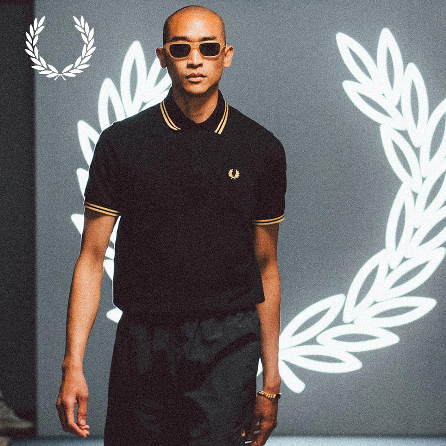 Ein Mann steht vor einer dunklen Wand mit Lorbeerkranz-Grafik. Er trägt ein schwarzes Polohemd und Sonnenbrille, modern und sportlich.
