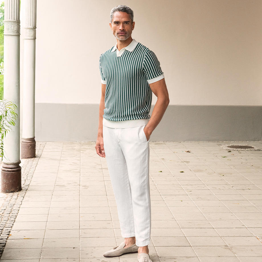 Ein Mann steht auf einer hellen Terrasse vor Säulen. Er trägt ein grünes, geripptes Poloshirt mit weißem Muster und eine weiße Hose mit hellen Mokassins.