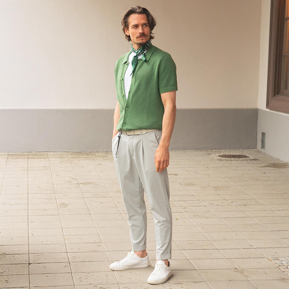 Ein Mann posiert im Halbschatten eines Durchgangs. Er trägt ein grünes Polohemd mit weißem Seidenschal, kombiniert mit einer hellen Hose und weißen Sneakers.