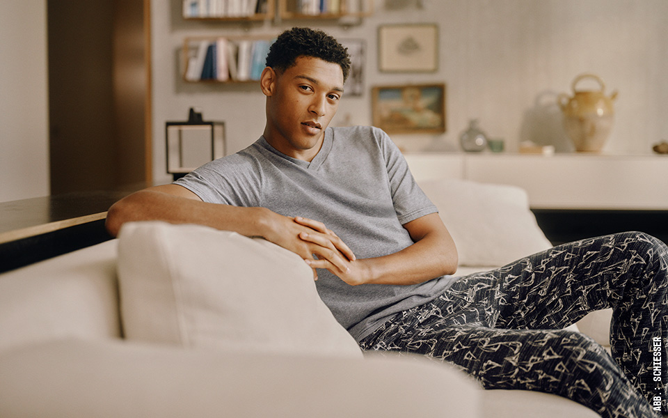 Ein junger Mann sitzt auf einem Sofa in einem Wohnzimmer. Er trägt einen Schiesser-Pyjama.