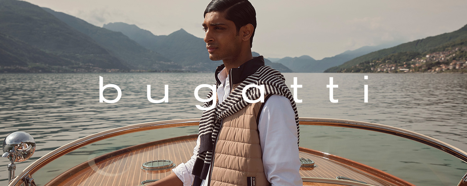 Mann in eleganter Freizeitkleidung von bugatti auf einem Holzmotorboot vor Bergkulisse: beige Steppweste, weißes Hemd, weißer Pullover mit schwarzen Streifen über den Schultern, weiße Hose – stilvolles Sommeroutfit am See.