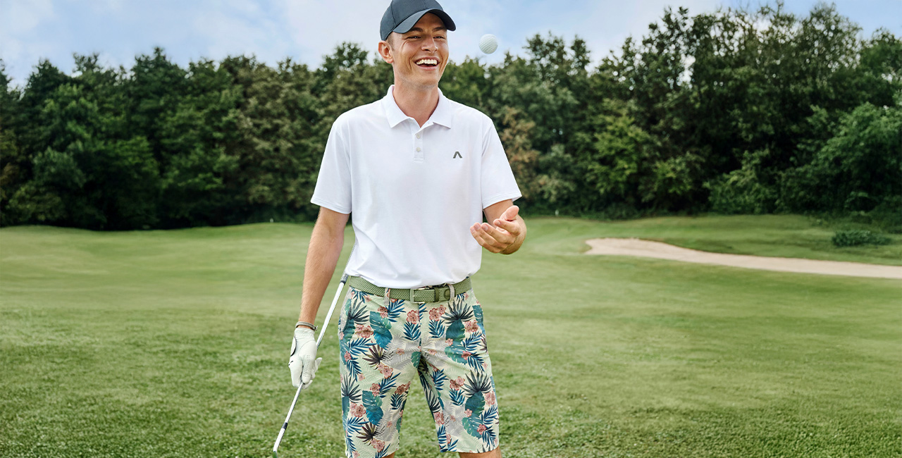 Ein Mann auf einem Golfplatz. Er trägt ein weißes Polo-Shirt, eine gemusterte Bermuda-Shorts und eine blaue Golfkappe.