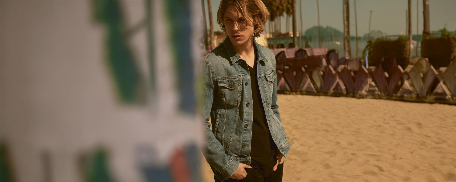Ein Mann in schwarzem T-Shirt, dunkler Hose und offener Jeansjacke steht am Strand und schaut zur Seite. Im Hintergrund ist ein Strand zu sehen.