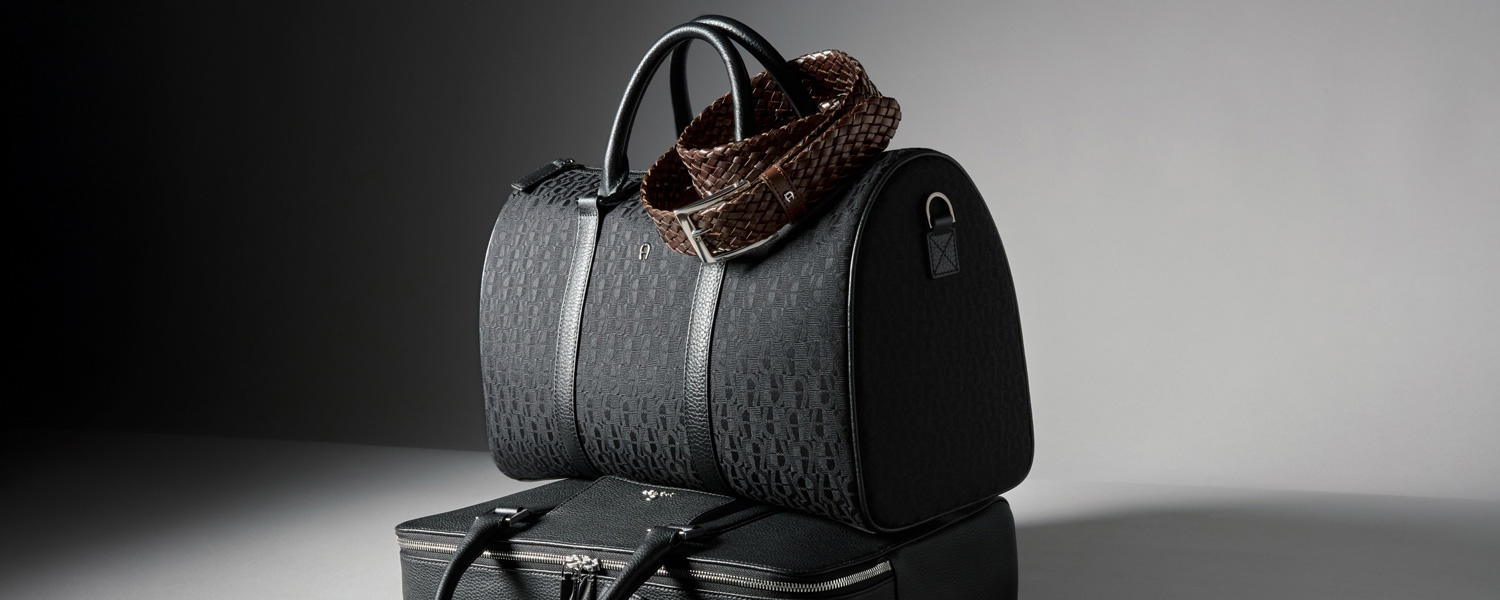 Zwei schwarze Taschen stehen übereinander vor einem grauen Hintergrund: eine klassische Aktentasche und ein Weekender, dessen Henkel mit einem braunen Gürtel umwickelt ist.