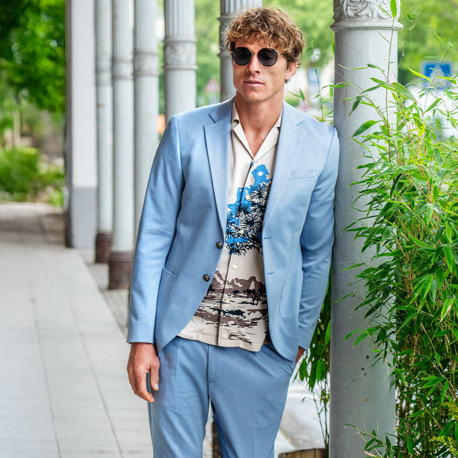 Ein Mann trägt einen hellblauen Anzug mit einem bedruckten Hemd und Sonnenbrille. Er steht lässig auf einem Weg mit Säulen und grüner Bepflanzung.