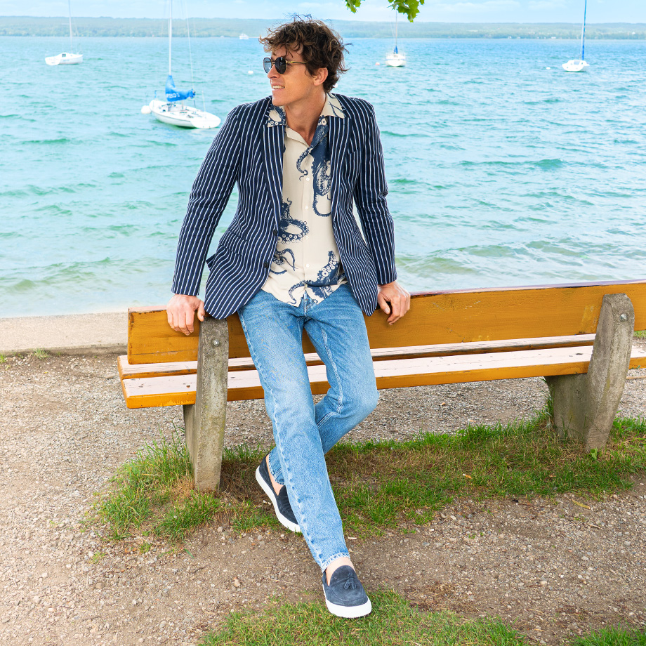 Mann in Jeans und gestreiftem Sakko sitzt entspannt auf einer Parkbank am See mit Segelbooten im Hintergrund.