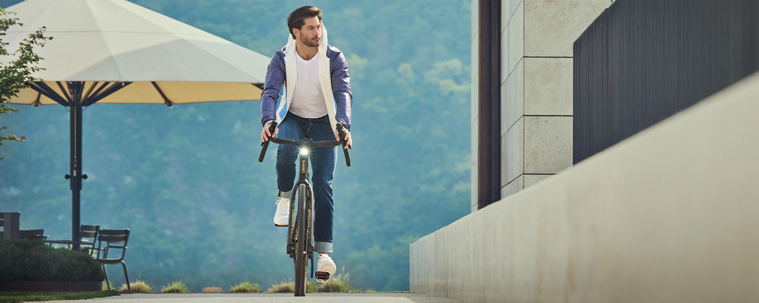 Ein Mann fährt bei strahlendem Wetter in einem lässigen Look – weißes T-Shirt, Jeans und offenes Hemd – auf einem Fahrrad. Im Hintergrund sind ein großer Sonnenschirm und grüne Natur zu sehen.