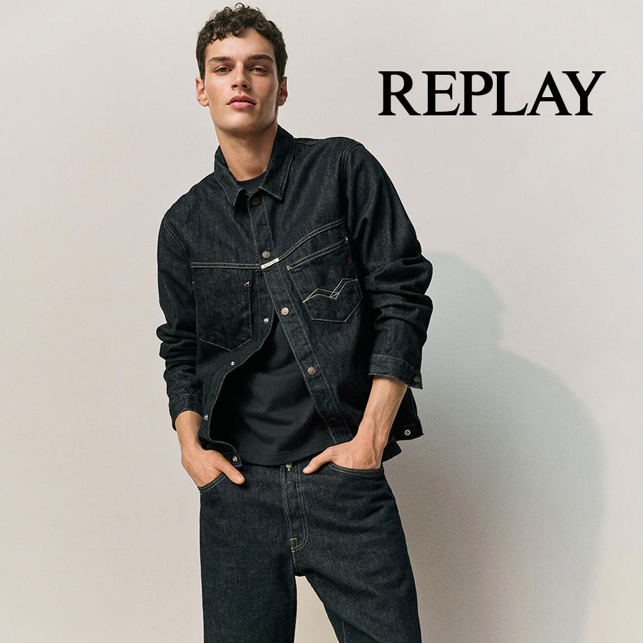 Junger Mann trägt ein dunkles Jeans-Outfit von Replay und lehnt locker an der Wand.