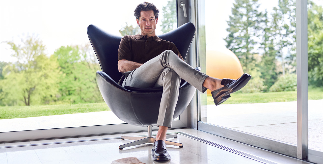 Mann in elegantem, modernem Outfit mit grauer Hose und braunem Polo sitzt entspannt in einem schwarzen Sessel vor einer großen Glasfront mit Blick ins Grüne.