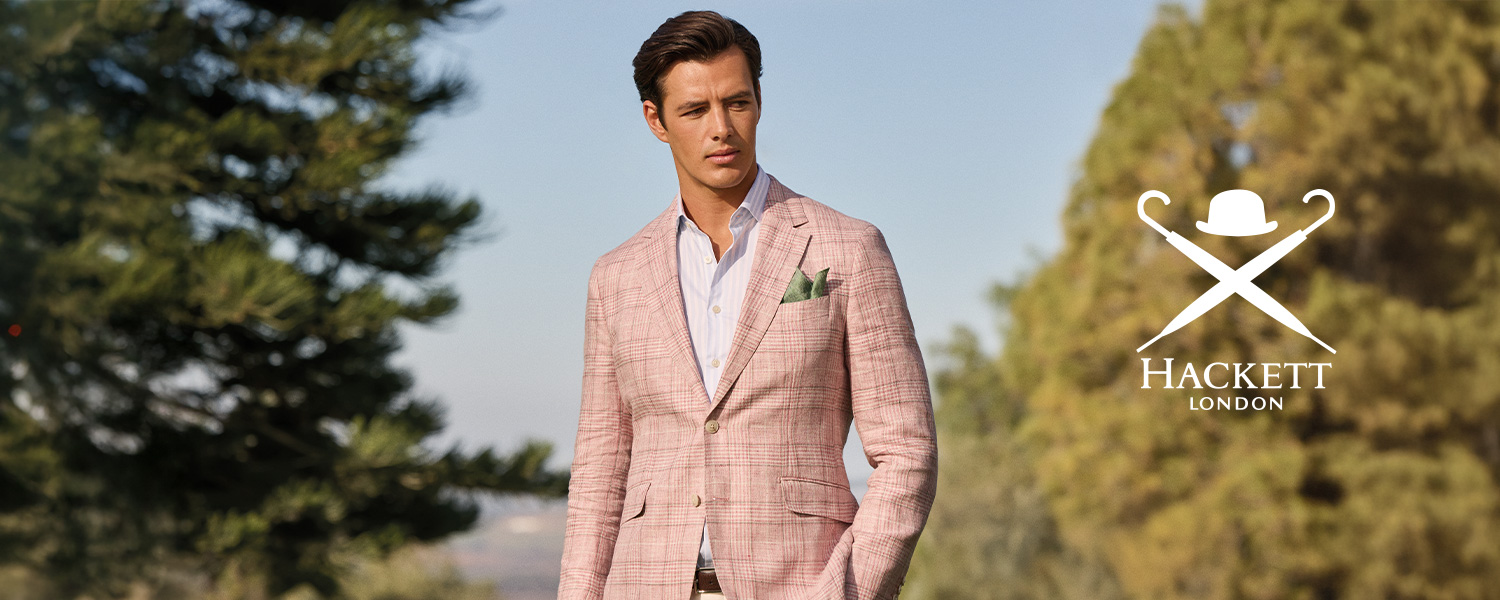 Ein Mann trägt ein elegantes, rosafarbenes kariertes Sakko mit weißem Hemd und heller Hose vor sommerlicher Kulisse.