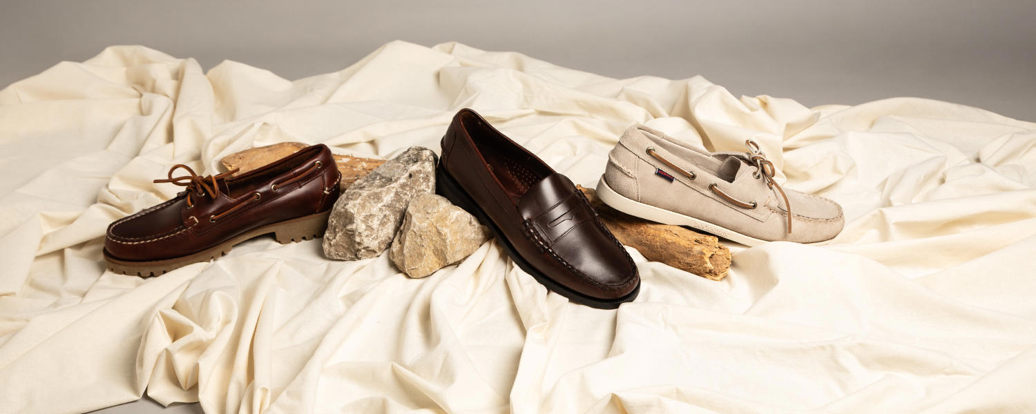 Zwei Bootsschuhe und 1 Loafer der Marke Sebago in Braun- und Beigetönen, dekorativ auf heller Stoffunterlage mit Naturmaterialien wie Steinen und Holz arrangiert.