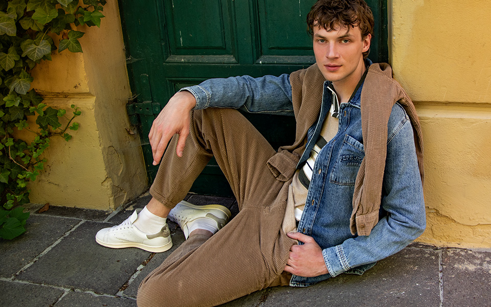 Ein junger Mann lehnt an einer alten Tür. Er trägt eine Cordhose, Poloshirt und eine Jeansjacke. Ein Cordhemd liegt über seinen Schultern