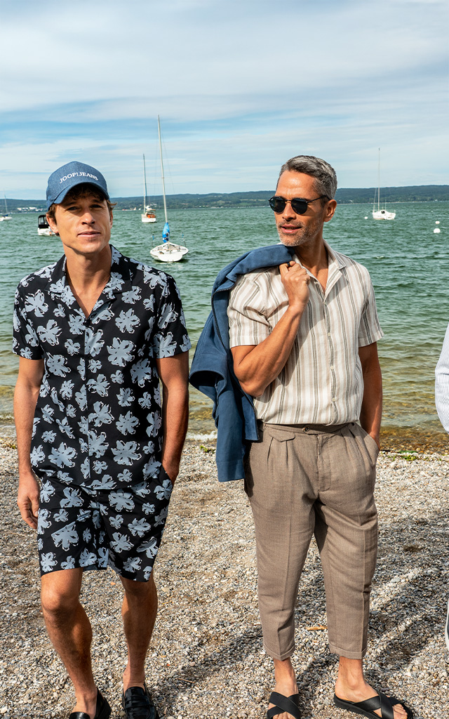 Drei Männer stehen nebeneinander am Strand. Sie tragen leichte, sommerliche Outfits in Beige, Blau und Weiß – ideal für einen Urlaub am Meer.