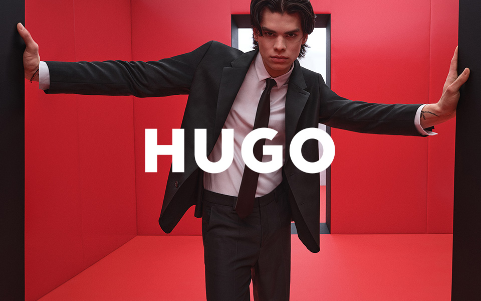 Ein junger Mann in einem roten Raum. Er trägt einen schwarzen HUGO-Anzug, mittiges HUGO-Logo