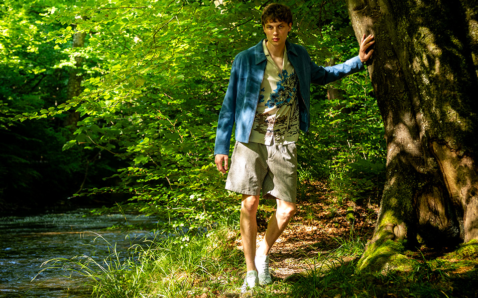 Ein junger Mann im Wald. Er trägt eine Lederjacke, ein Hemd und eine Shorts