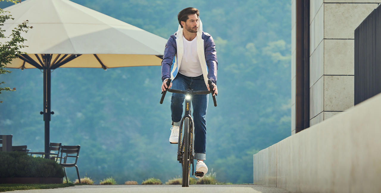 Ein Mann in Jeans, weißem T-Shirt und blauer Jacke fährt auf einem modernen Fahrrad vor einem Gebäude mit heller Steinfassade; im Hintergrund stehen Gartenmöbel und ein großer Sonnenschirm vor bewaldeter Berglandschaft.