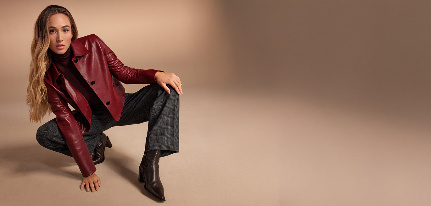 Eine junge Frau im Studio mit beigem Background. Sie trägt eine rote Lederjacke von MILESTONE und kniet auf dem Boden.