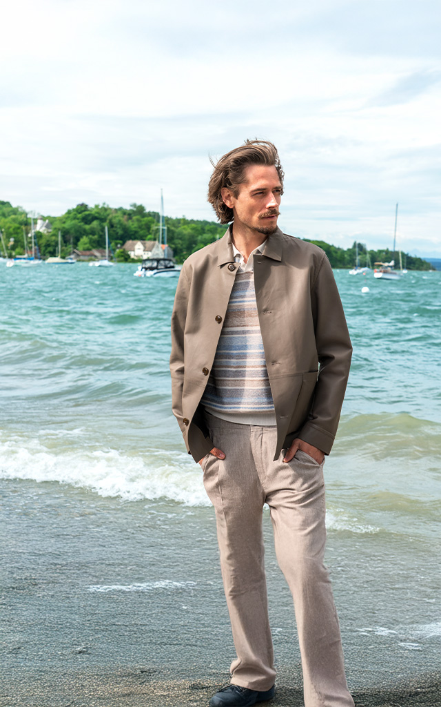 Ein Mann mit beiger Lederjacke und beige-blau-weiß gestreiftem Polo-Shirt steht entspannt am Seeufer mit Segelbooten im Hintergrund.