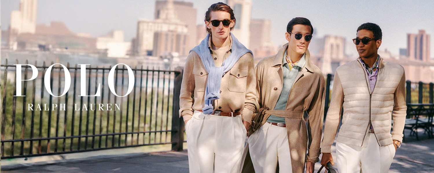 Drei stilvoll gekleidete Männer in hellen Trenchcoats, Jacken und Overshirts kombiniert mit hellen Hosen und lässigen Freizeitlooks spazieren nebeneinander auf einem Uferweg – urban, modern und elegant.