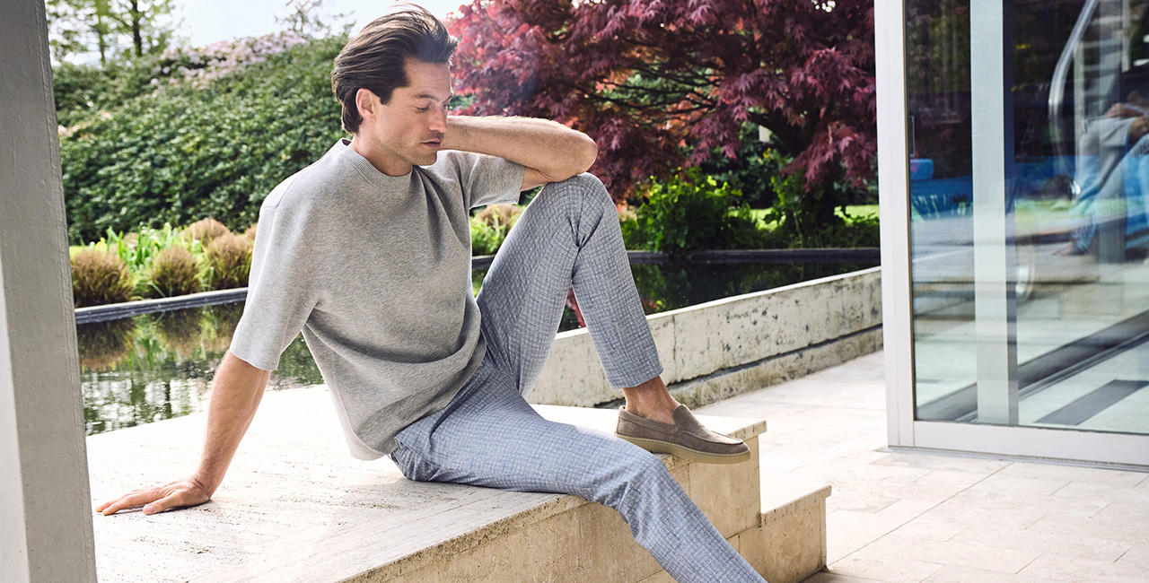 Alt Text Jersey: Ein stilvoll gekleideter Mann sitzt entspannt auf einer Steintreppe vor moderner Architektur, trägt graues Kurzarmshirt und karierte Stoffhose – sommerlich-urbaner Freizeitlook.