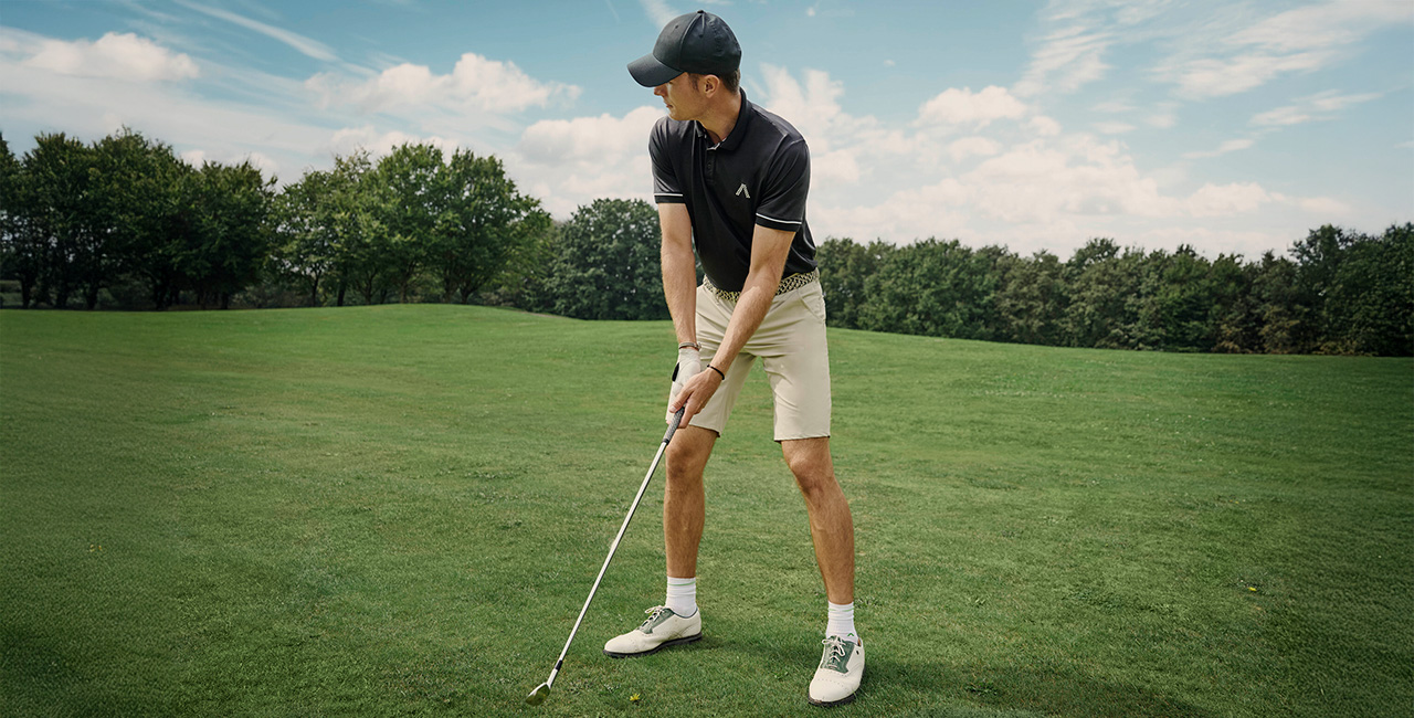 Mann in schwarzem Poloshirt und beigen Shorts bereitet sich auf dem Golfplatz auf einen Schlag vor – sportlich-eleganter Freizeitstil