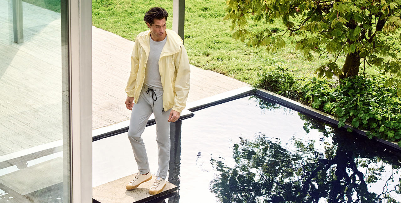 Mann in hellgrauem T-Shirt und Jogpant mit gelber Jacke steht an einem Wasserbecken vor einem modernen Gebäude – minimalistischer Sommerlook in natürlicher Umgebung.